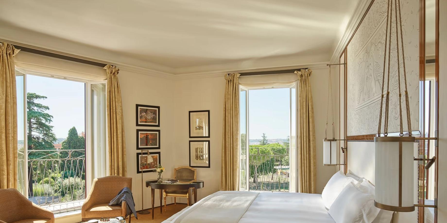A bedroom with pale stone walls, tall windows overlooking Rome, and natural light illuminating the bed