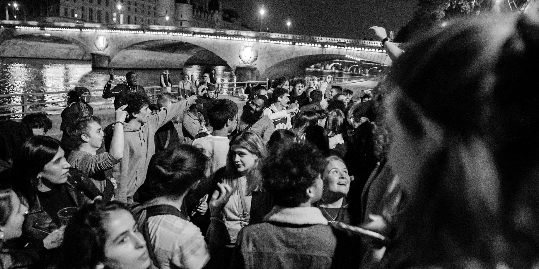 A night crowd celebrating on a Seine quai in Paris, a lit bridge behind, in black and white