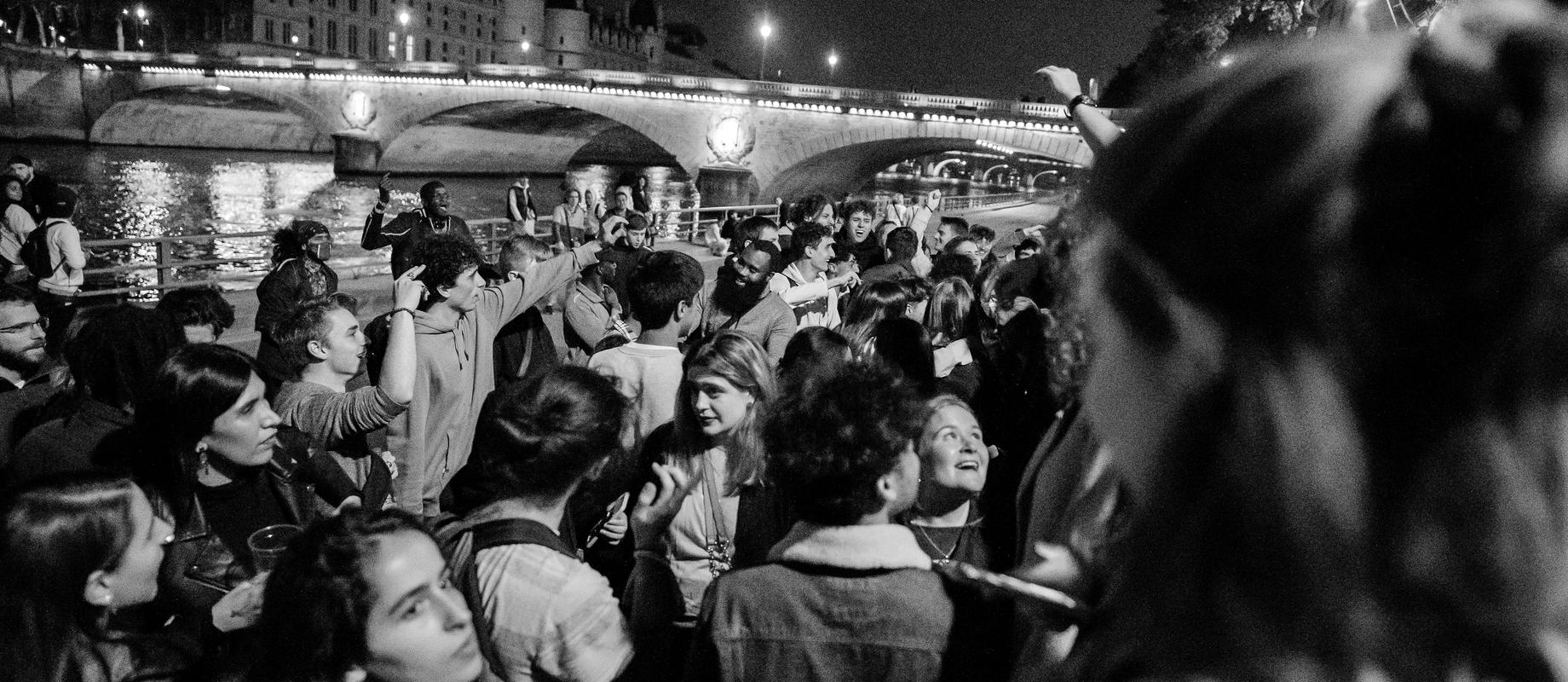 A night crowd celebrating on a Seine quai in Paris, a lit bridge behind, in black and white