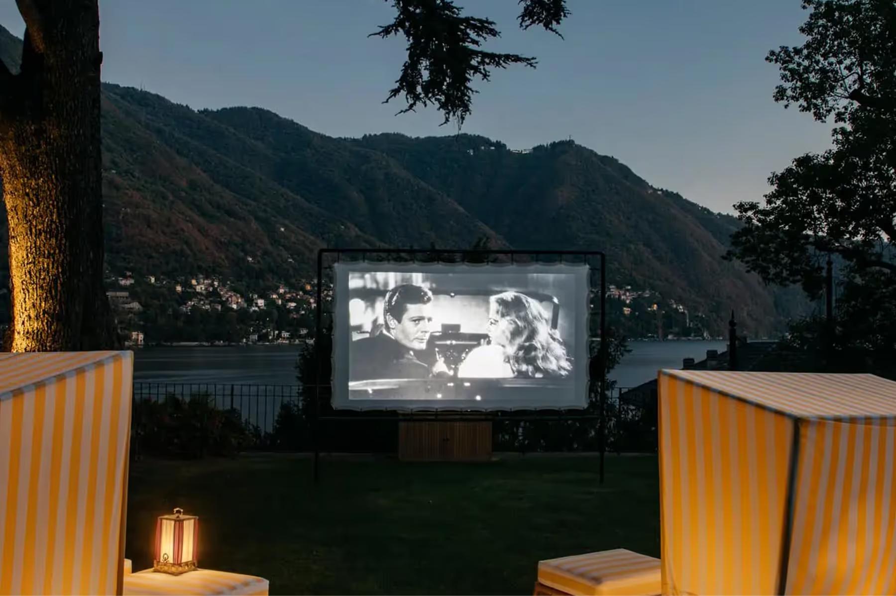 An outdoor movie screen glowing at dusk with upholstered seating and lake view