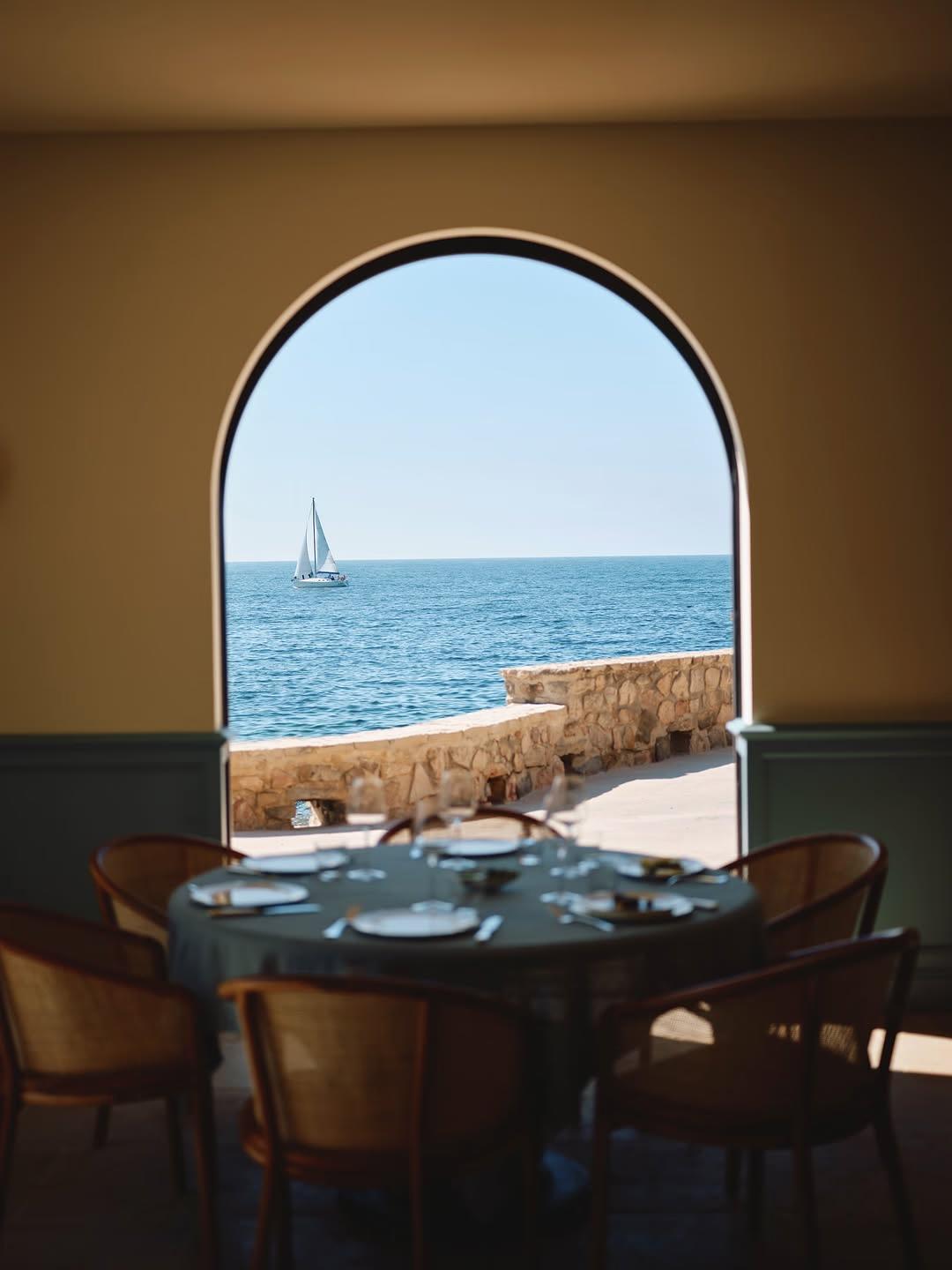 A table set for dinner frames a stone archway looking out to a passing sailboat