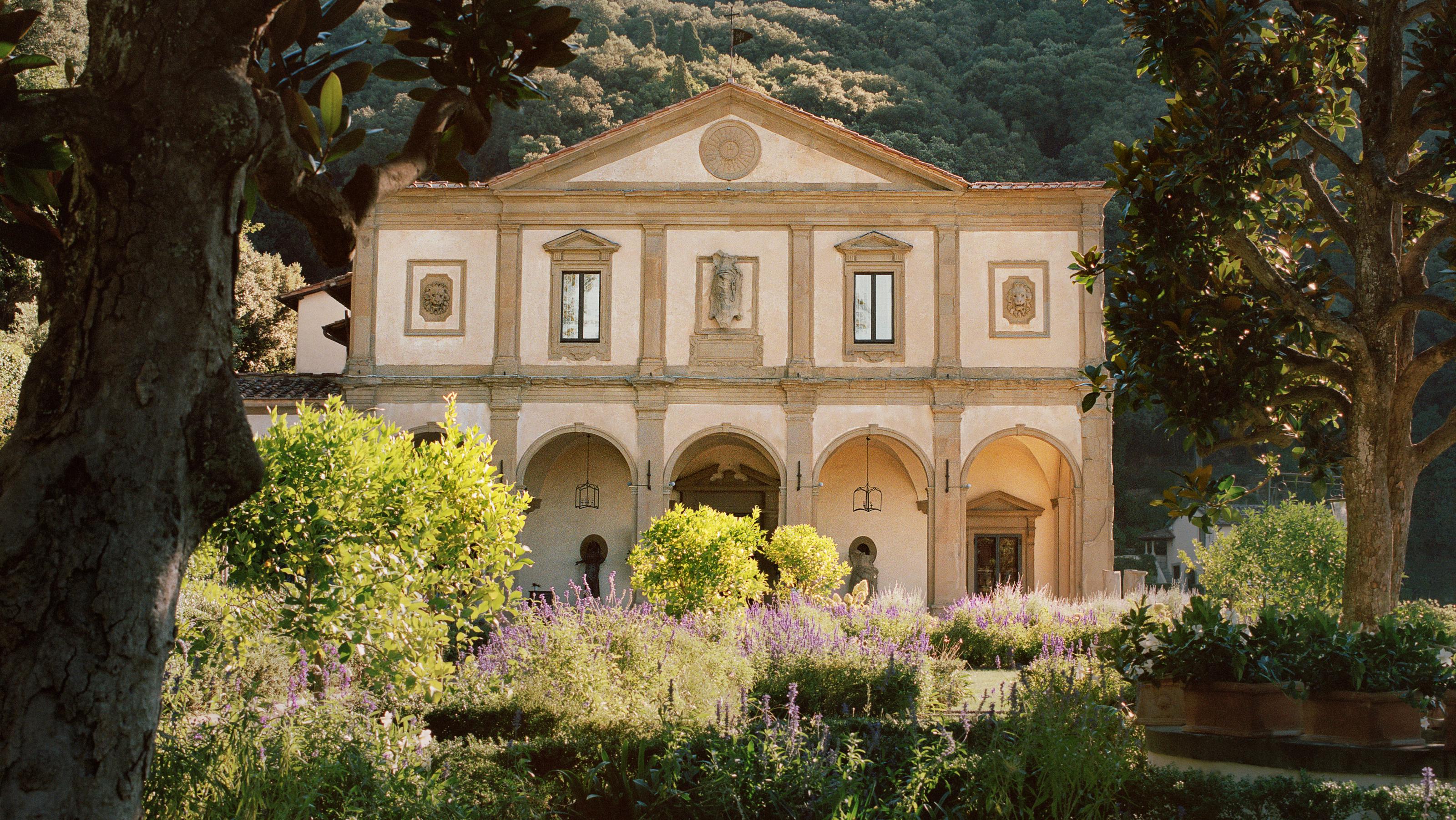 The terracotta-hued facade of the former monastery surrounded by cypress trees and lush gardens