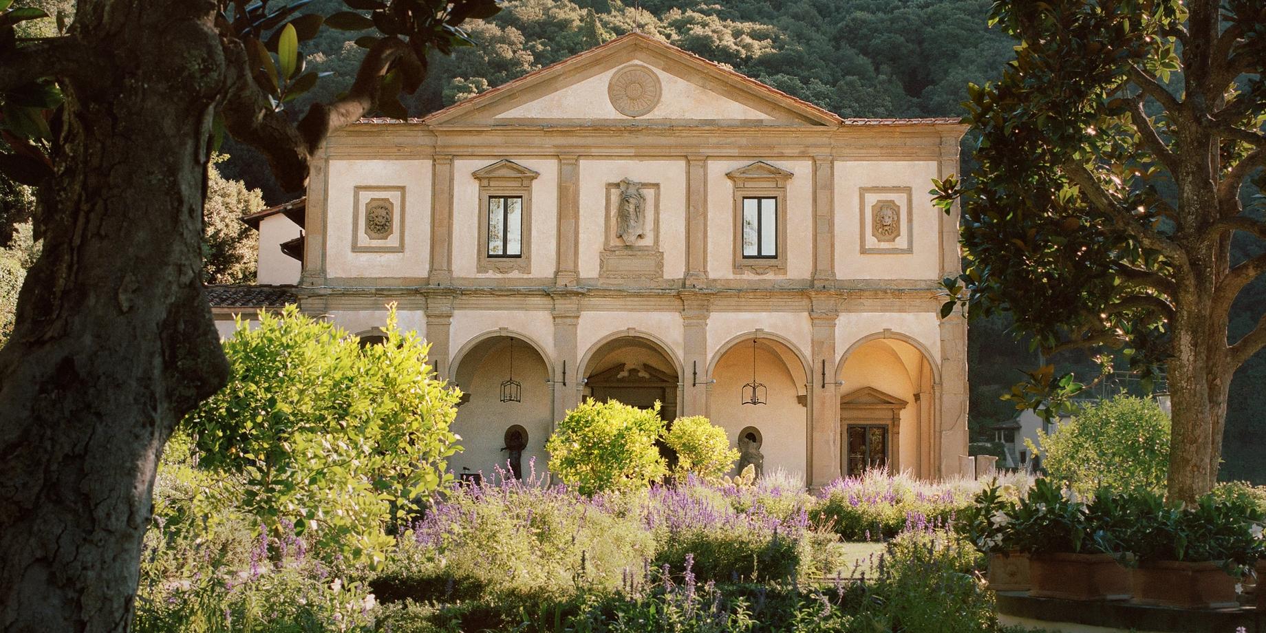 The terracotta-hued facade of the former monastery surrounded by cypress trees and lush gardens