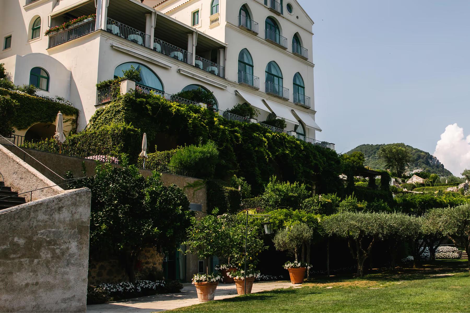 Caruso: white palazzo exterior with manicured evergreen hedges and terracotta potted plants on sloping lawn