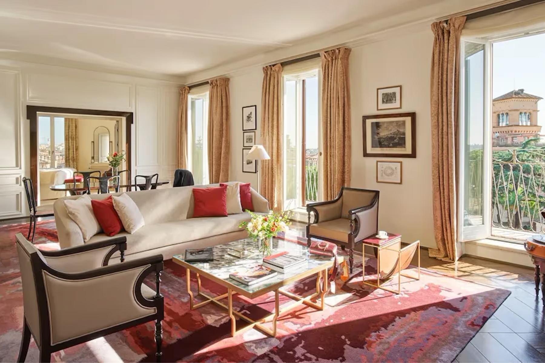 Hotel Eden: a living room with cream furnishings, red accent chairs, decorative artwork, and tall windows overlooking Rome