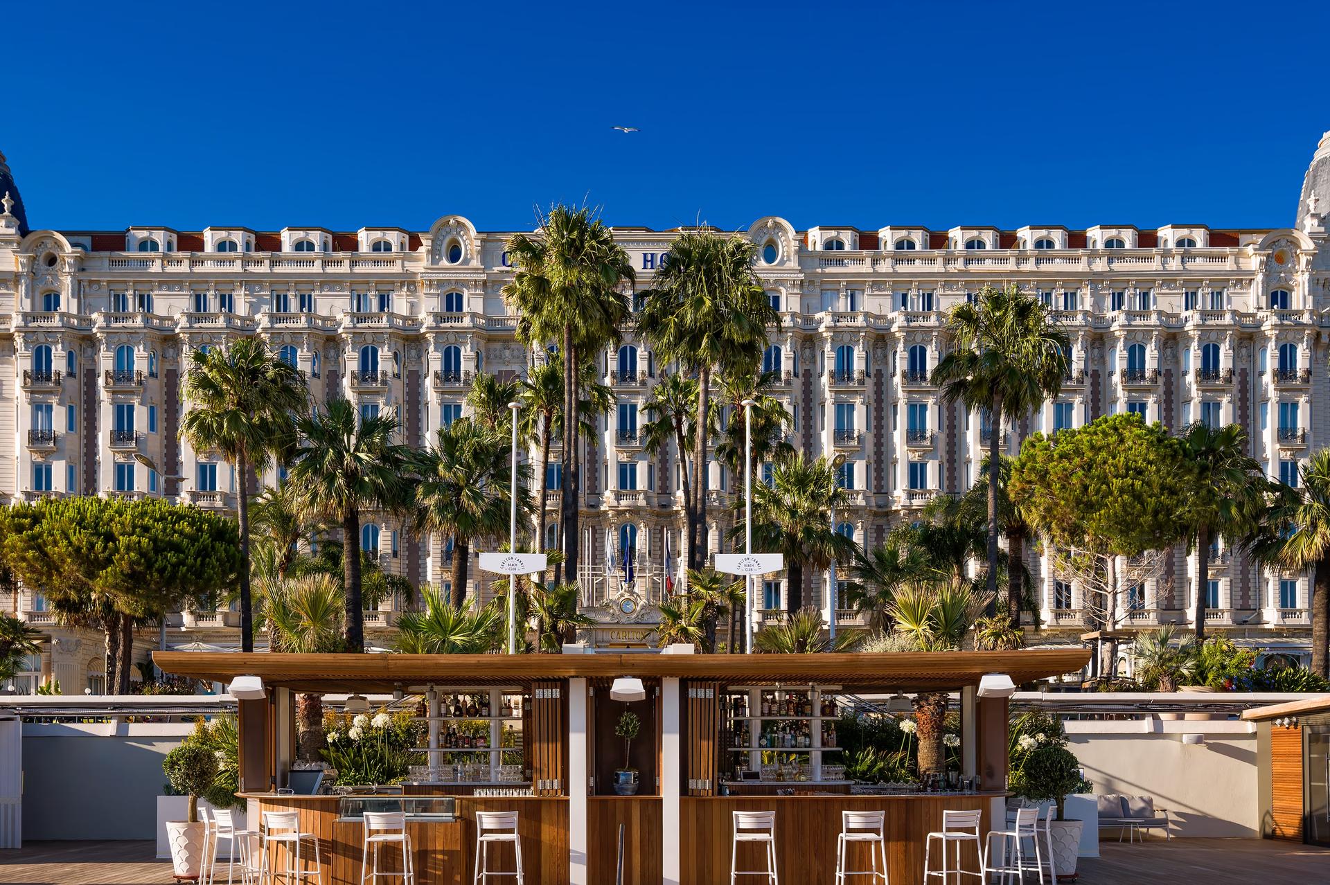 Front facade of Carlton Cannes overlooking the Mediterranean with palm-lined beach and turquoise water
