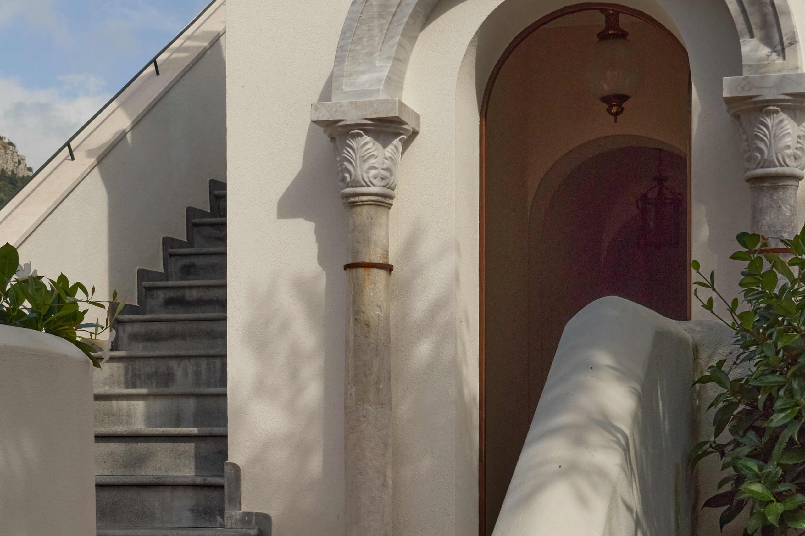 Caruso: outdoor archway with ornate column, coastal stairs, and crimson wooden door on whitewashed building