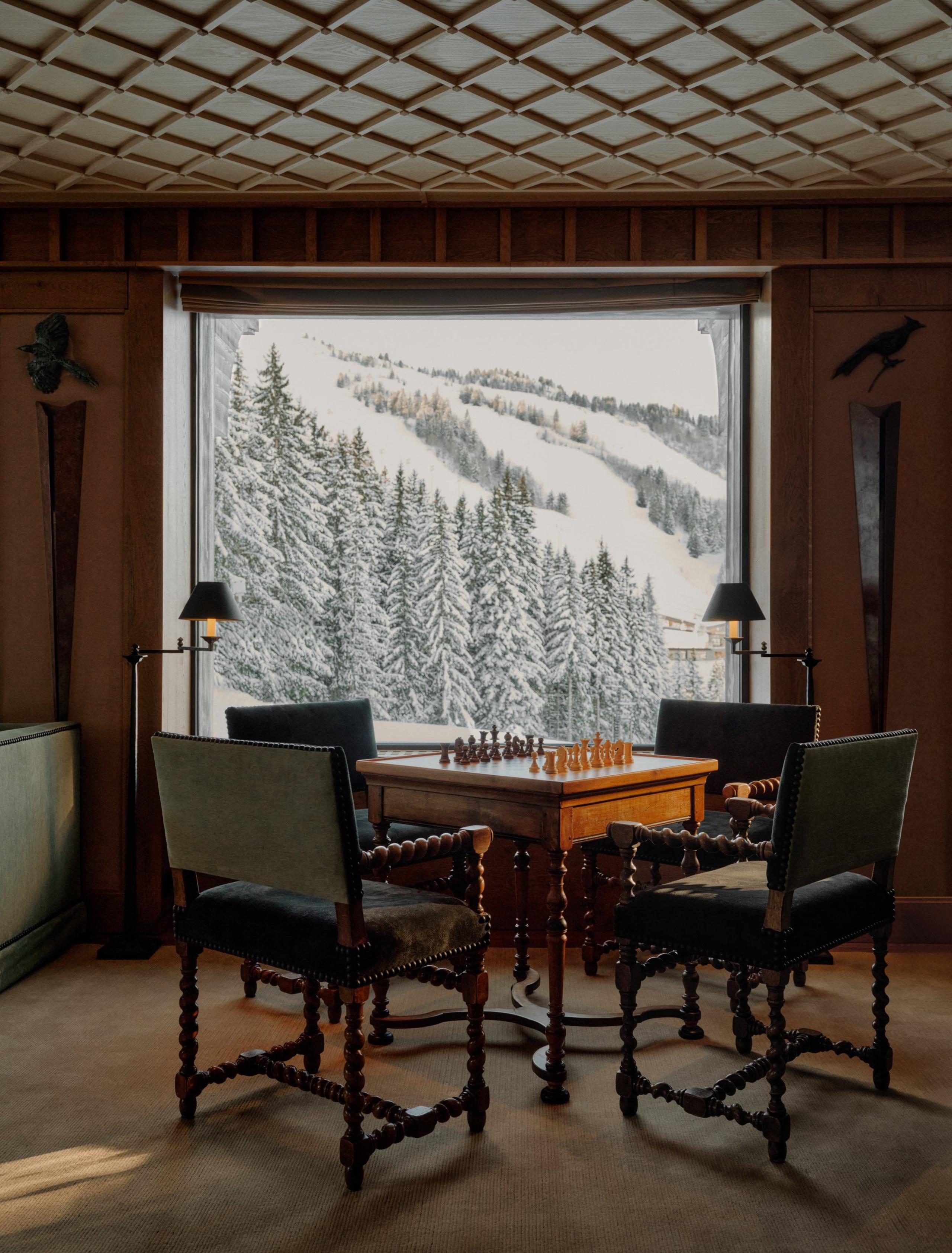 Chess table surrounded by four chairs beneath a coffered ceiling, facing a window over the piste