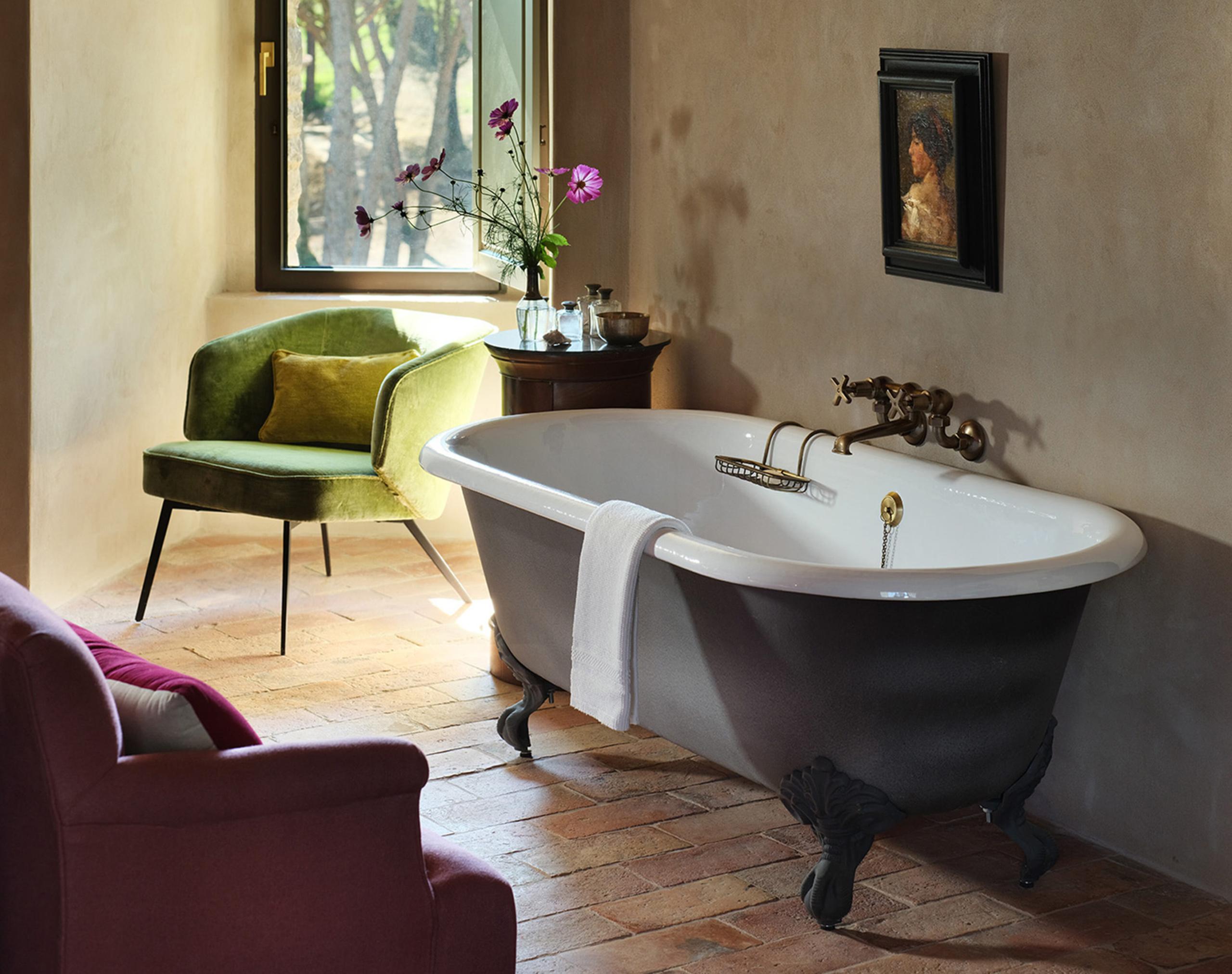 Castello di Reschio: a bathroom with a white clawfoot tub beside an arched window, an olive velvet armchair, and terracotta tile