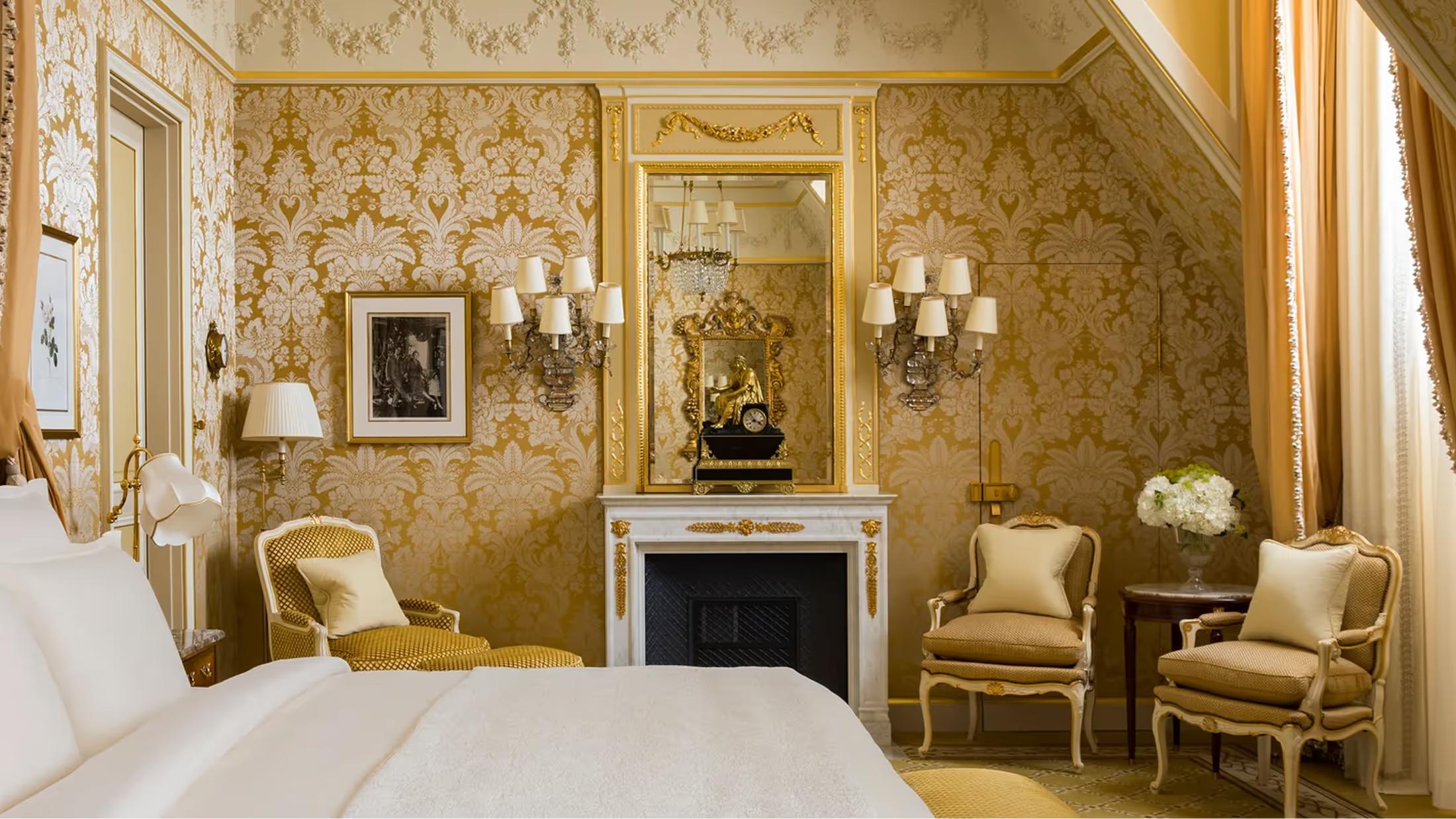 A Belle Époque suite with gold damask walls, cream bedding, fireplace mantel, and gilt-framed mirror