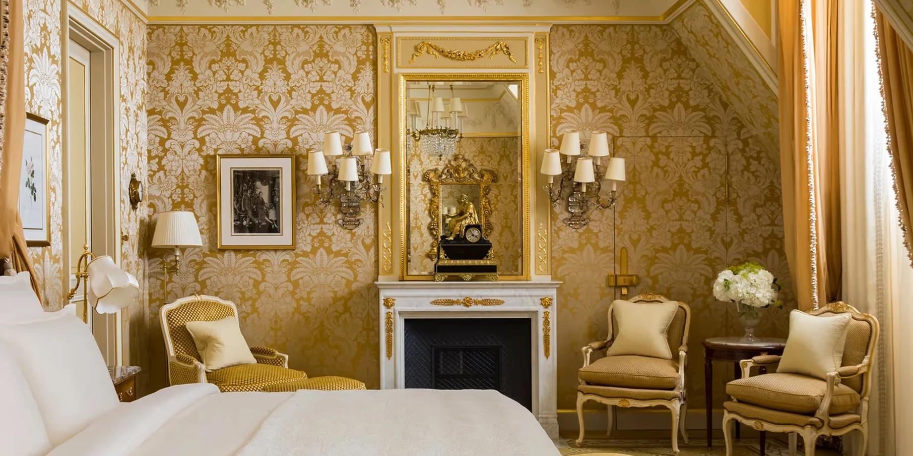 A Belle Époque suite with gold damask walls, cream bedding, fireplace mantel, and gilt-framed mirror