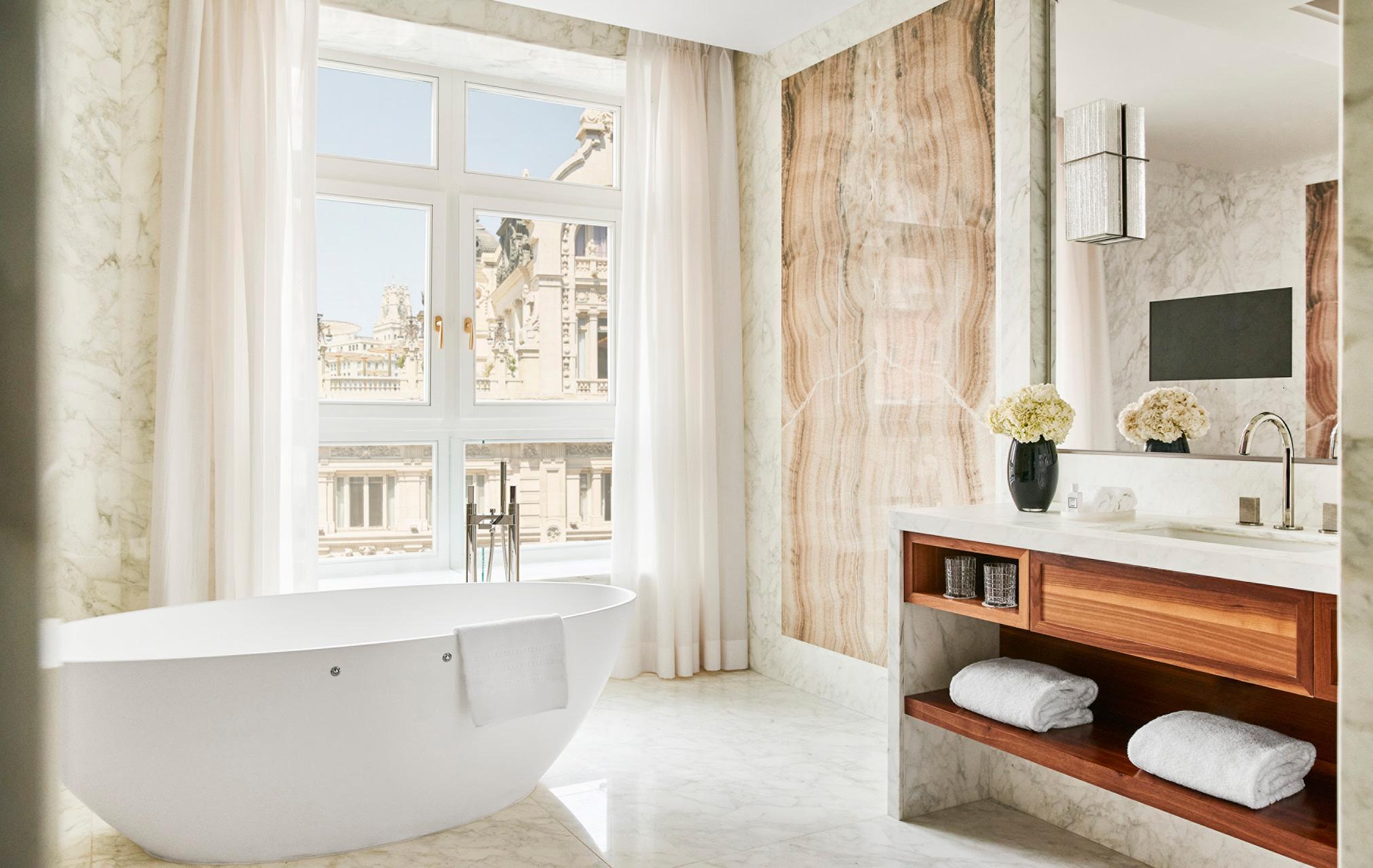 Four Seasons Hotel Madrid: spa bathroom with freestanding tub, marble surround, wood vanity, and arched window with city views