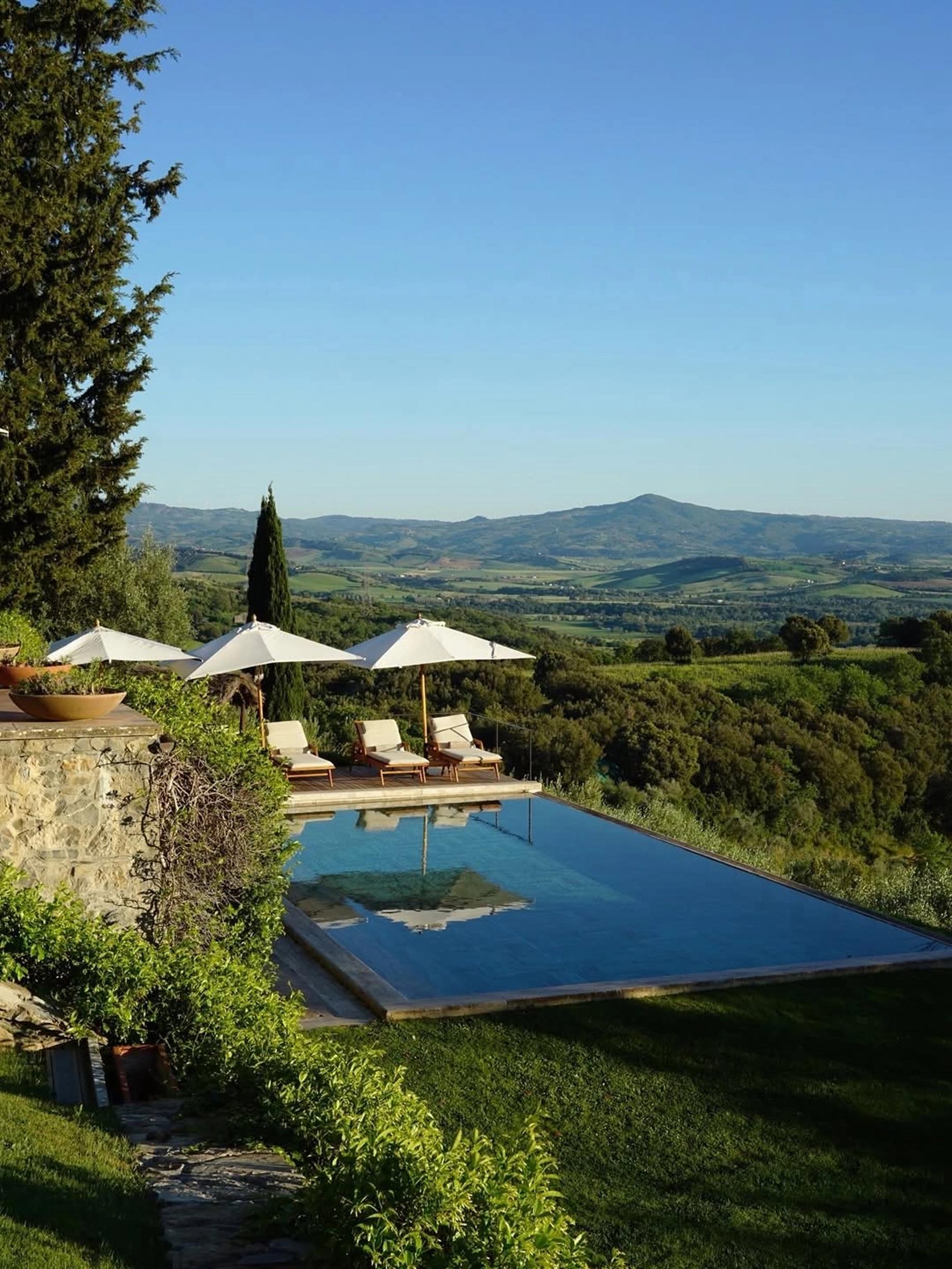 A pool with white umbrellas and sun loungers on a hillside, green valleys and a mountain beyond
