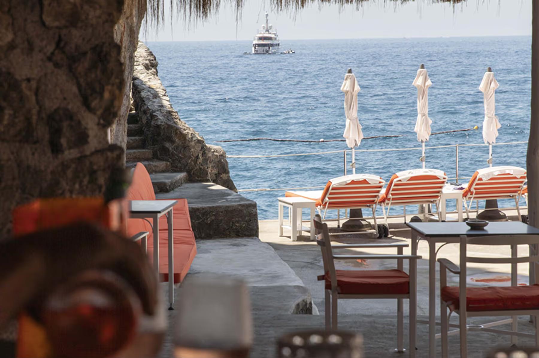 A cliffside restaurant terrace with striped umbrellas, open to the sea and mountains