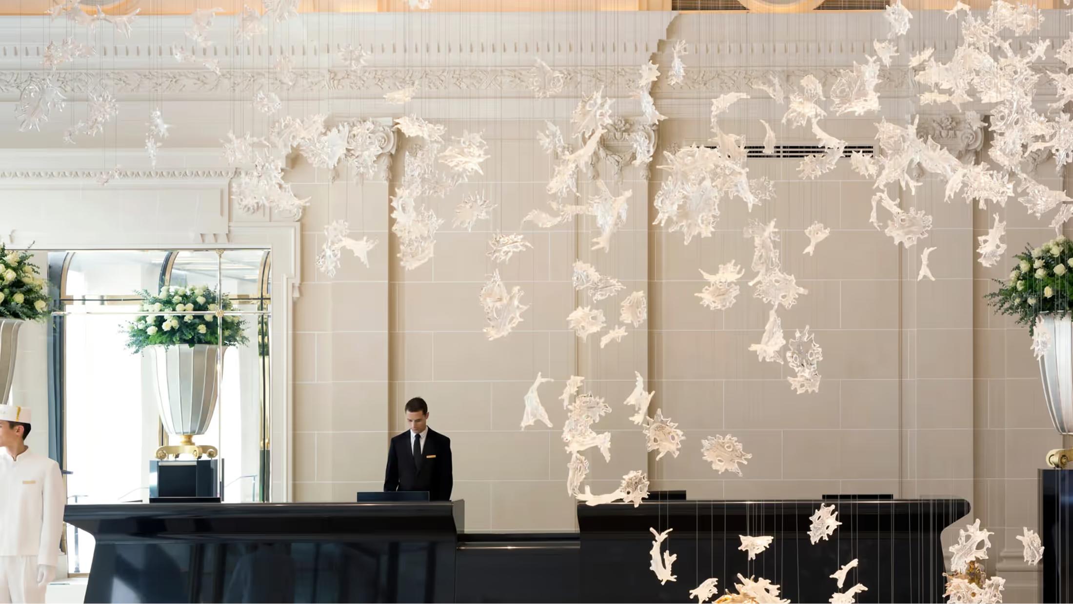 A refined Parisian lobby with cream-colored walls, suspended floral installations, and a concierge at a dark desk