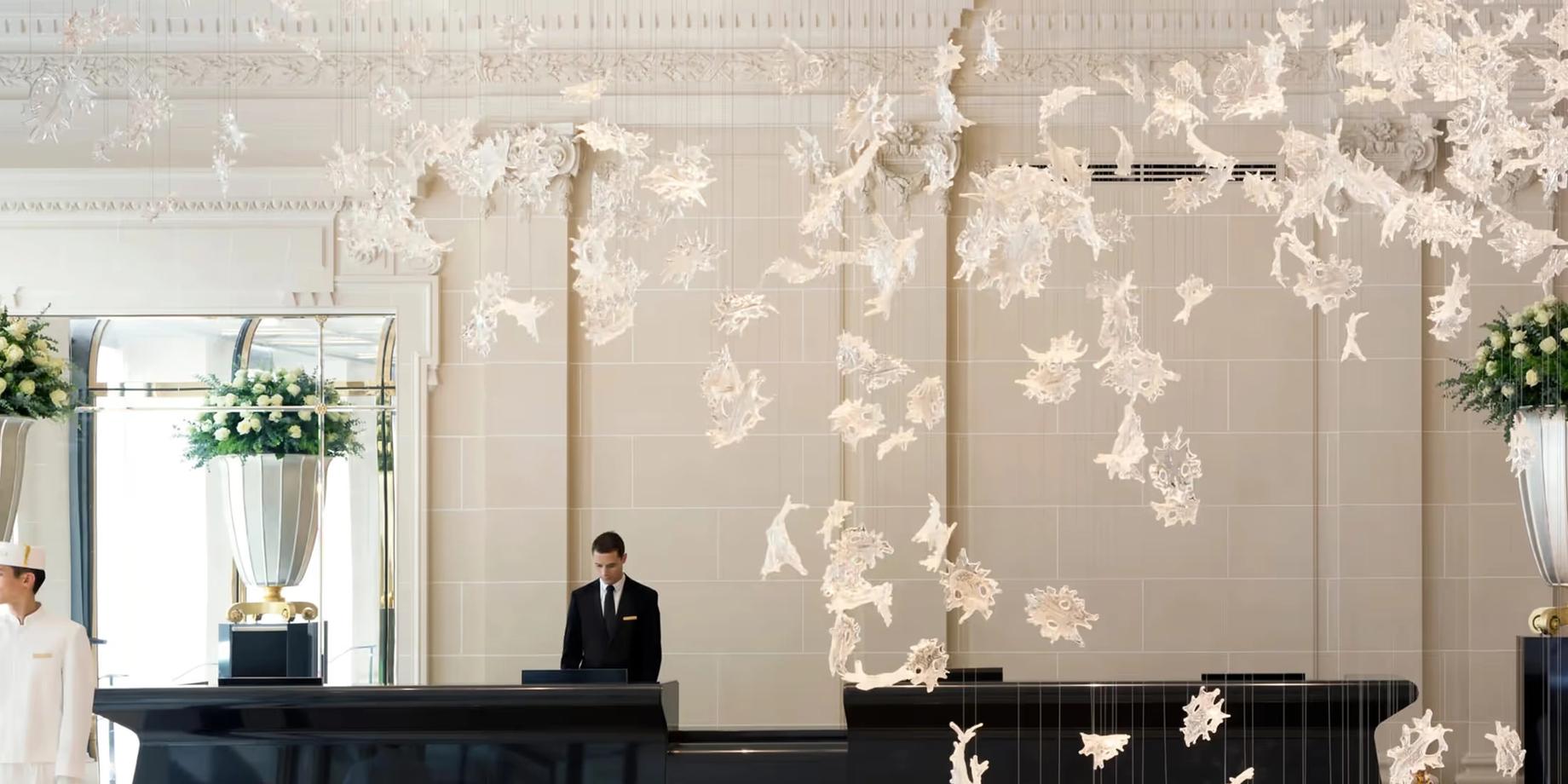 A refined Parisian lobby with cream-colored walls, suspended floral installations, and a concierge at a dark desk