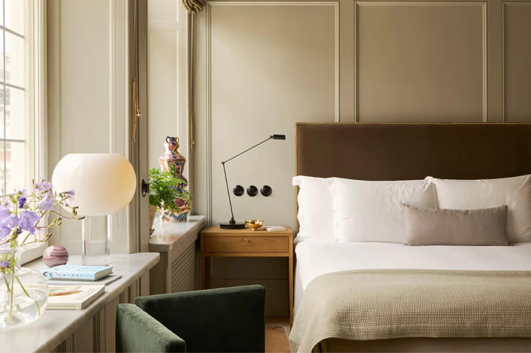 A bedroom with tan headboard, white pillows, glass bedside table, and flowers in a vase by the window