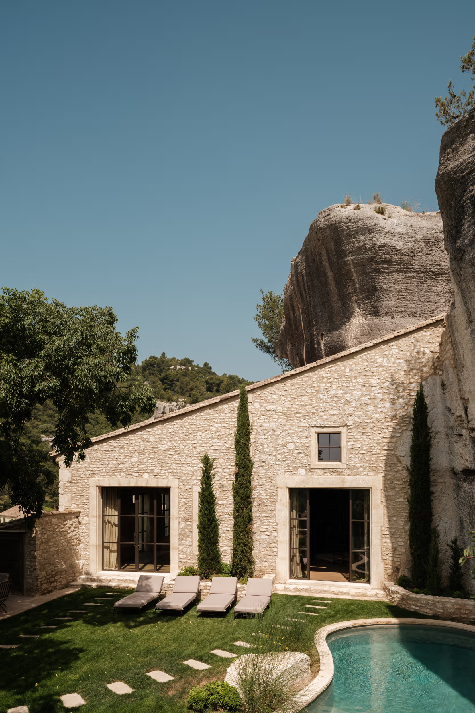 La Bergerie: stone courtyard with pale loungers, turquoise pool, raw limestone cliff wall, cypress wind-break privacy
