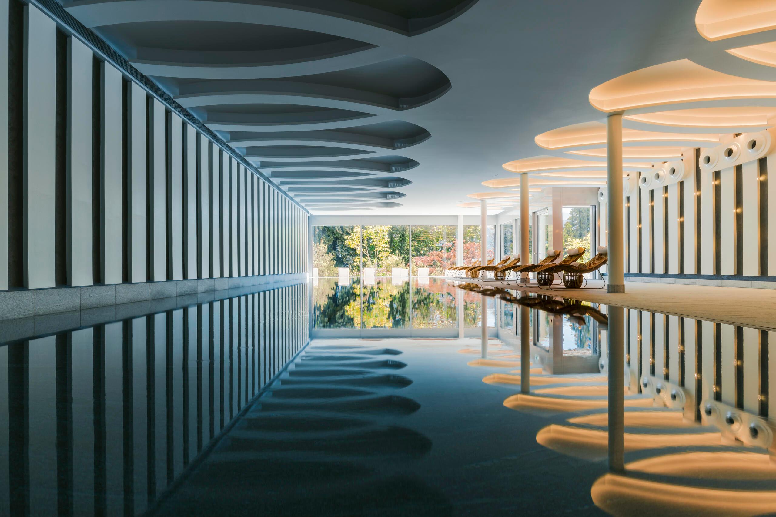 Modern spa pool with reflective dark floor, white and teal striped ceiling panels, warm lighting, and lake views through glass