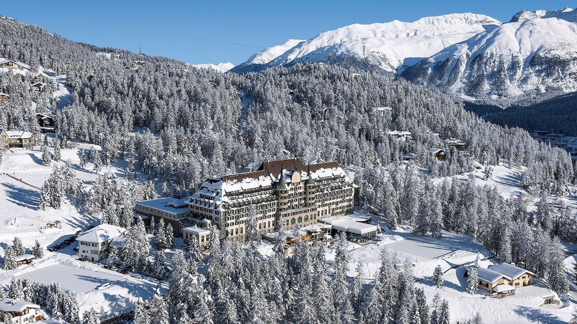 The hotel on a forested alpine plateau with snow-covered peaks in the distance