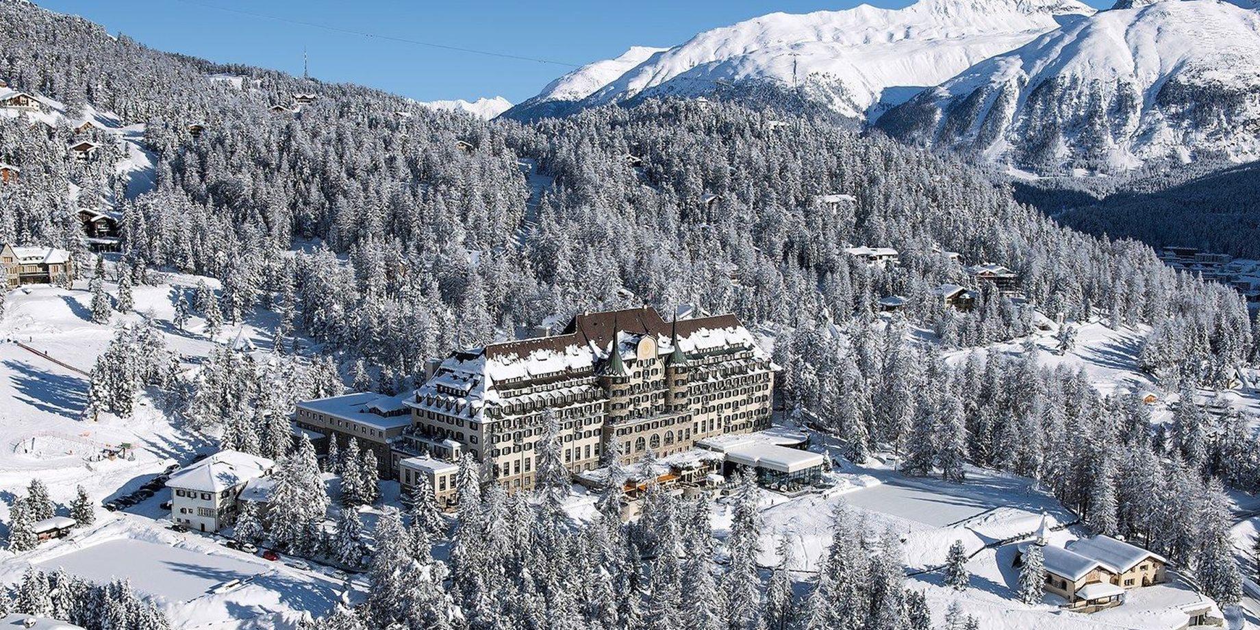The hotel on a forested alpine plateau with snow-covered peaks in the distance