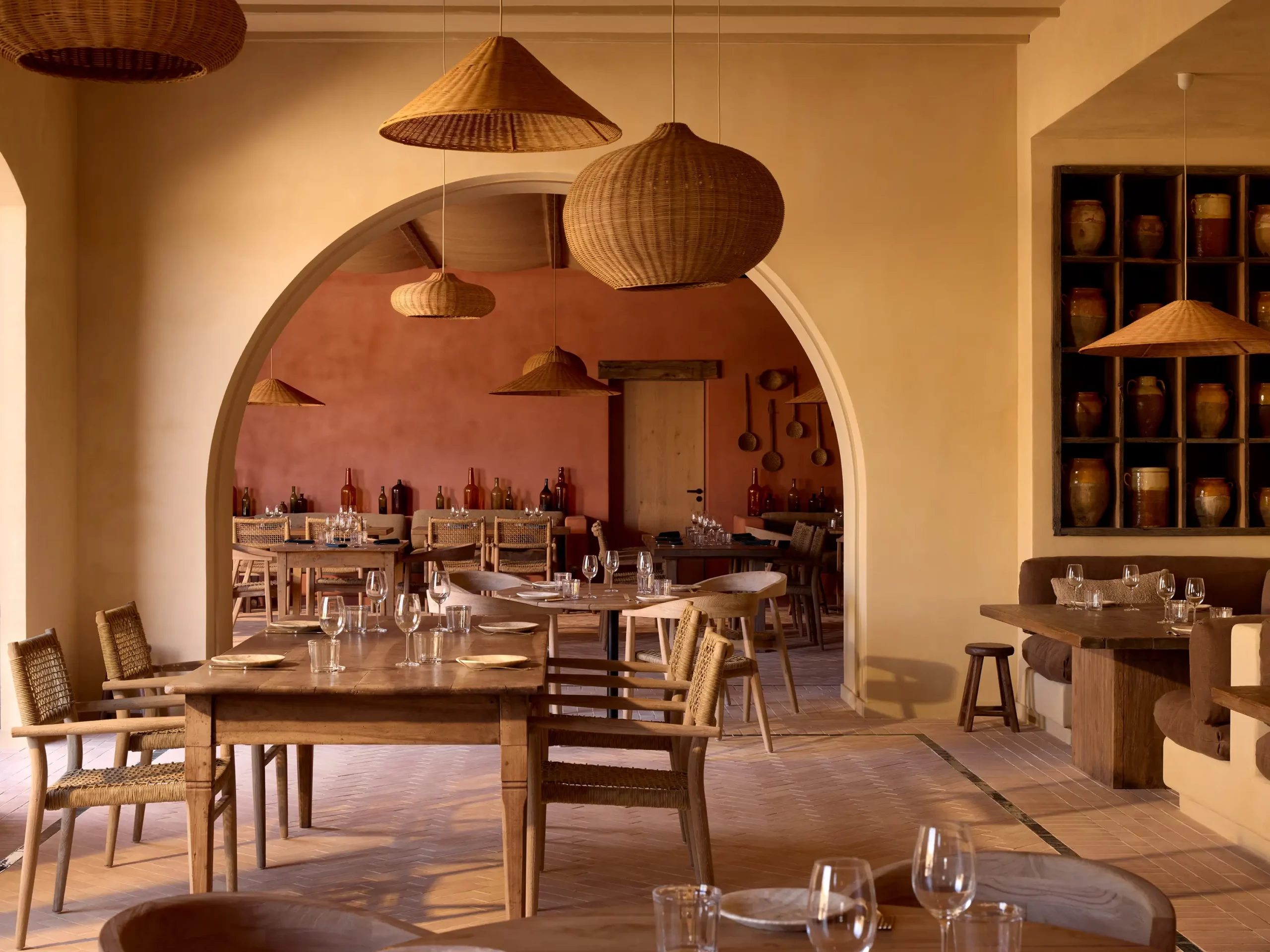 Woven rattan pendants hang over a rustic dining room with a terracotta arch and wine bottles