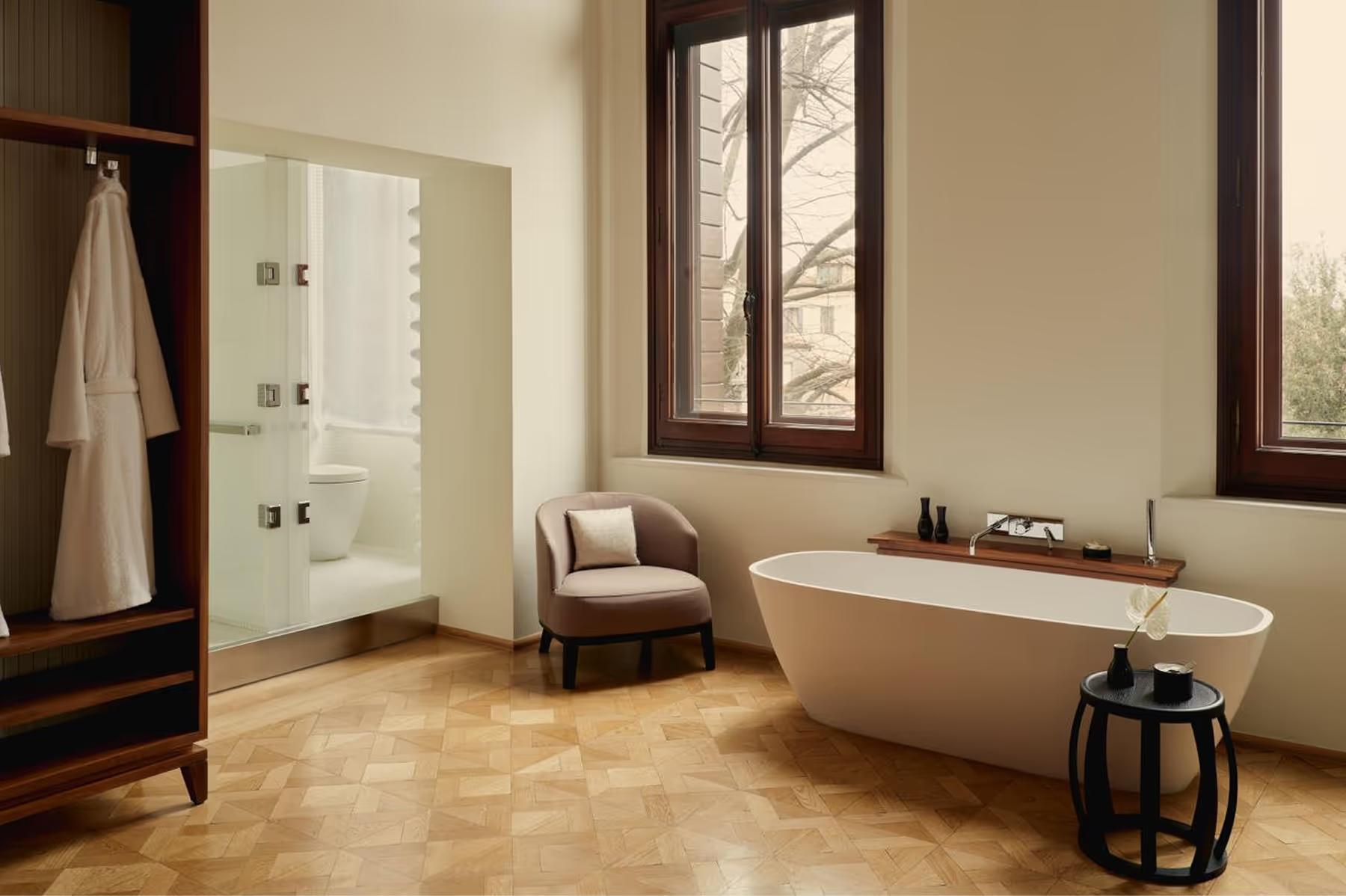 Aman Venice: a spa bathroom with a freestanding white stone tub, glass-enclosed shower, and windows opening to green views