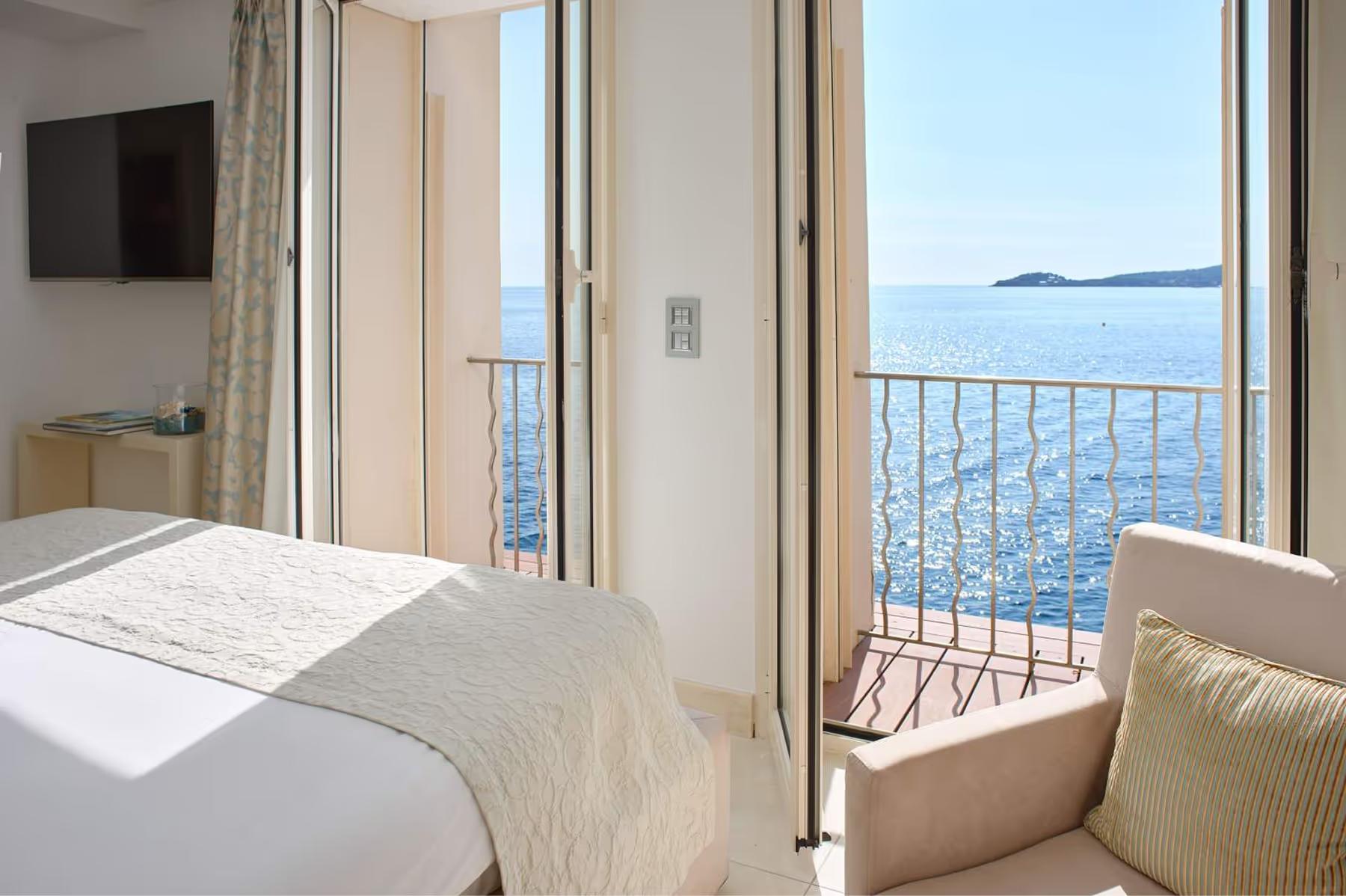 A bedroom with open glass doors to a balcony overlooking blue water