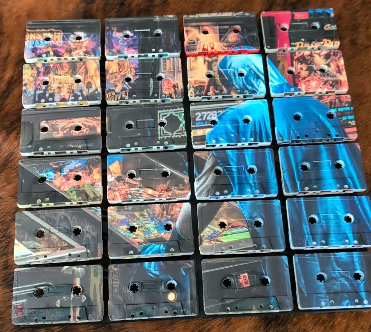 Twenty-four printed cassette faces in four columns and six rows, with busy pinball imagery on the left and blue clothing crossing several tapes on the right, resting on a brown-and-white hide surface.
