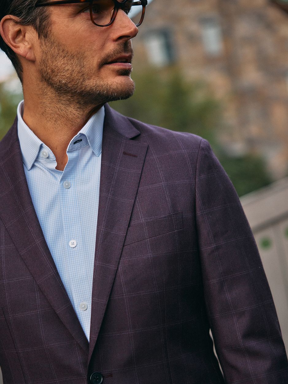 A close-up portrait of a man wearing eyeglasses, a light blue micro-grid dress shirt, and a dark plum windowpane blazer.