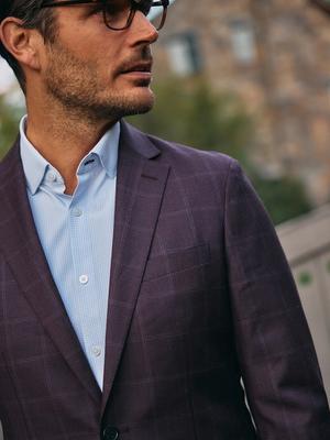A close-up portrait of a man wearing eyeglasses, a light blue micro-grid dress shirt, and a dark plum windowpane blazer.