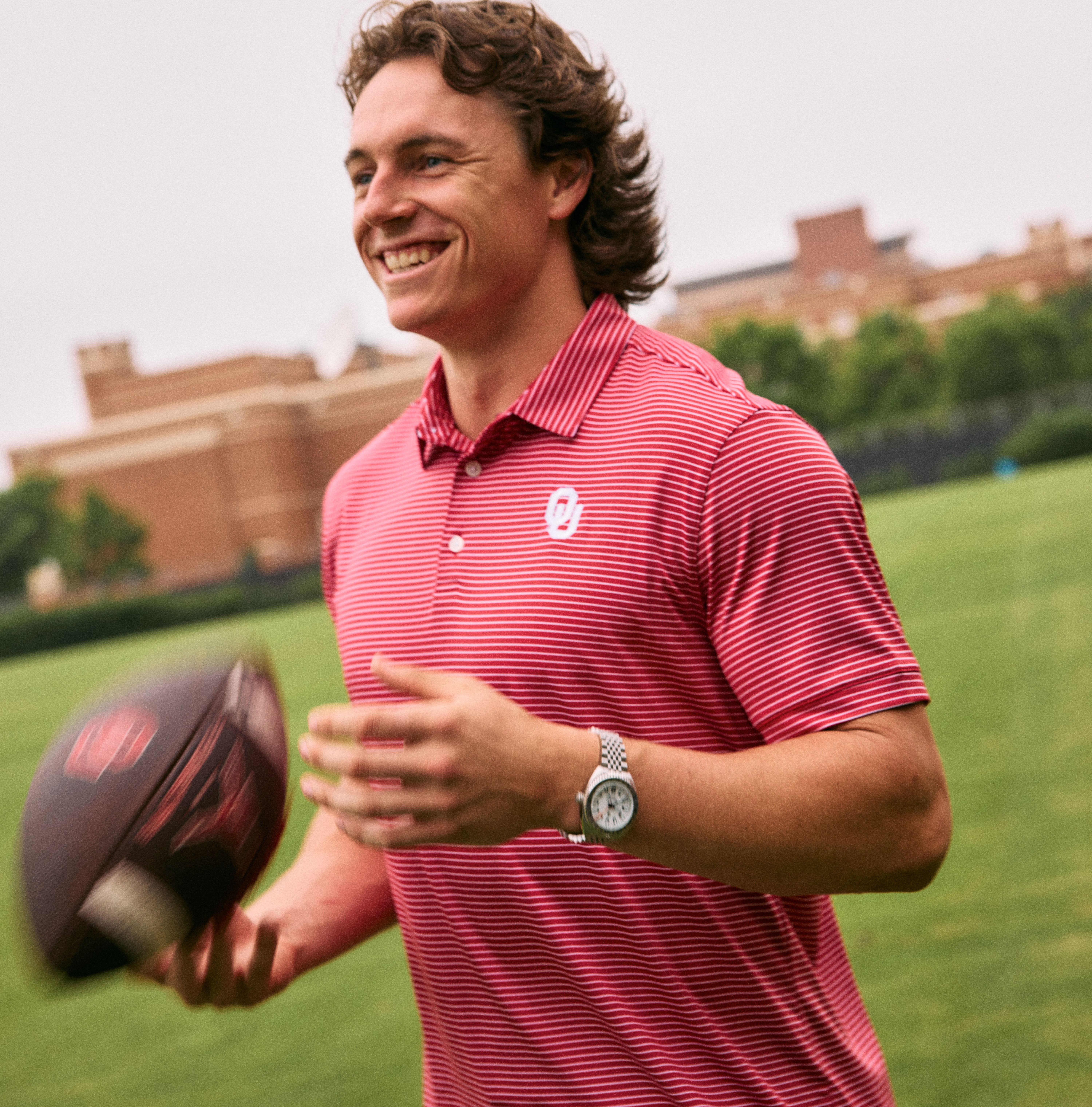 A man in a red and white micro-striped Oklahoma polo shirt smiling on an outdoor grassy field while catching a football.