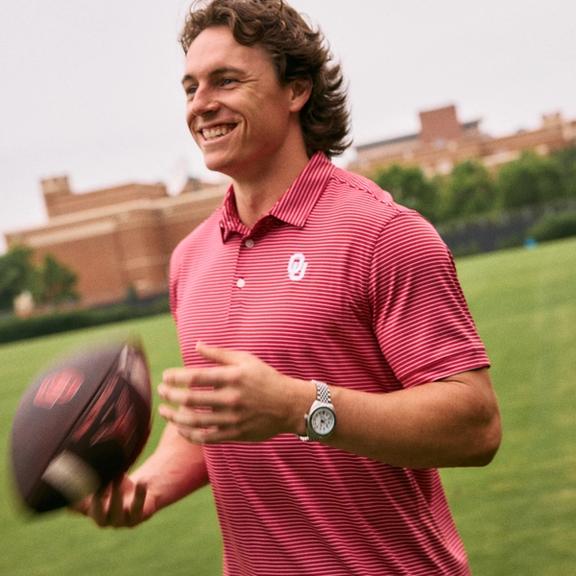A man in a red and white micro-striped Oklahoma polo shirt smiling on an outdoor grassy field while catching a football.