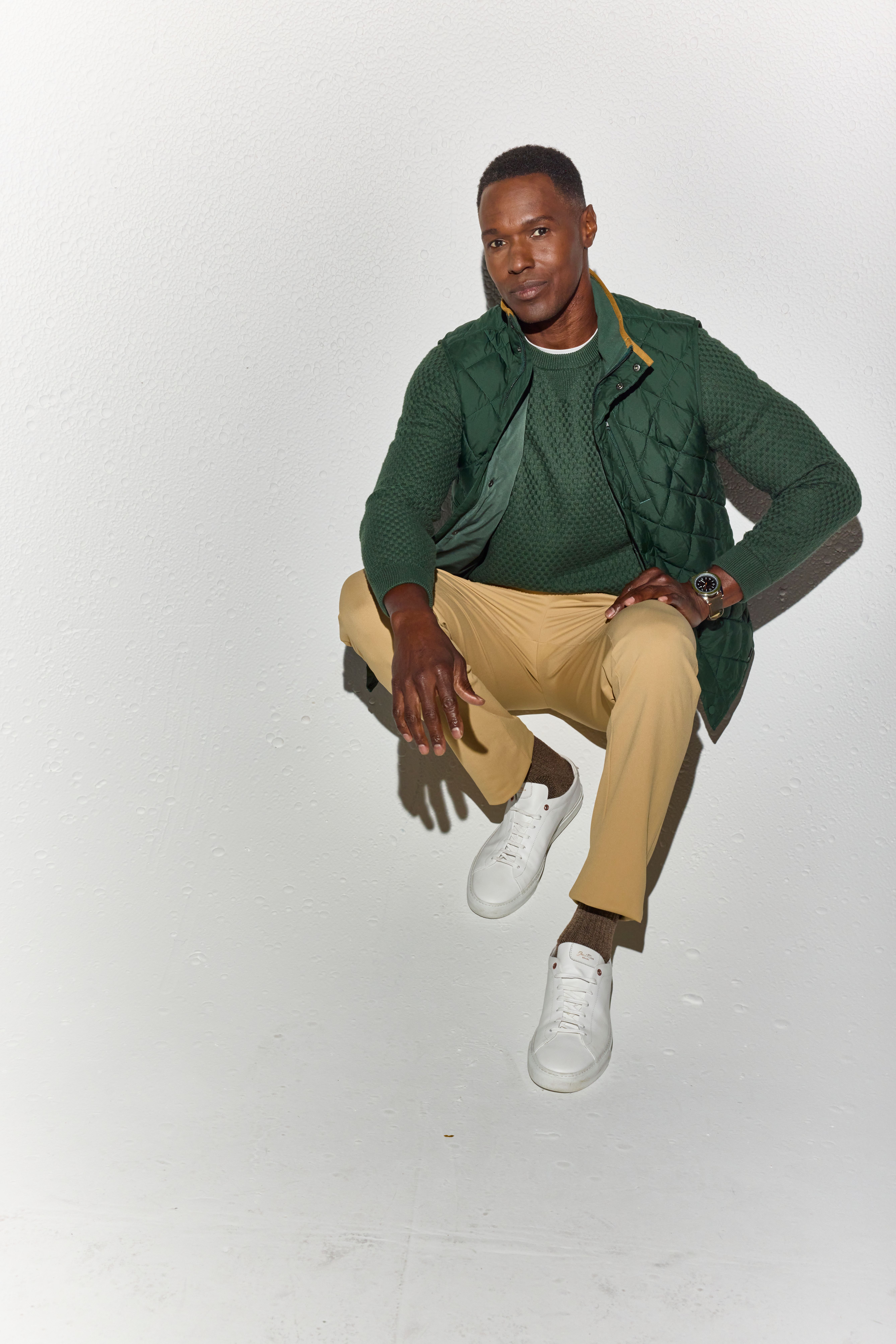 Man crouching in a green Hillcrest sweater layered under a quilted vest, with khaki pants