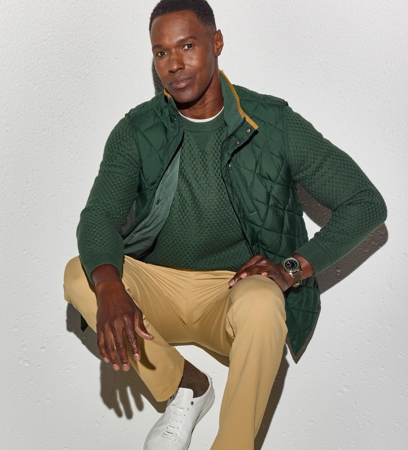 Man crouching in a green Hillcrest sweater layered under a quilted vest, with khaki pants