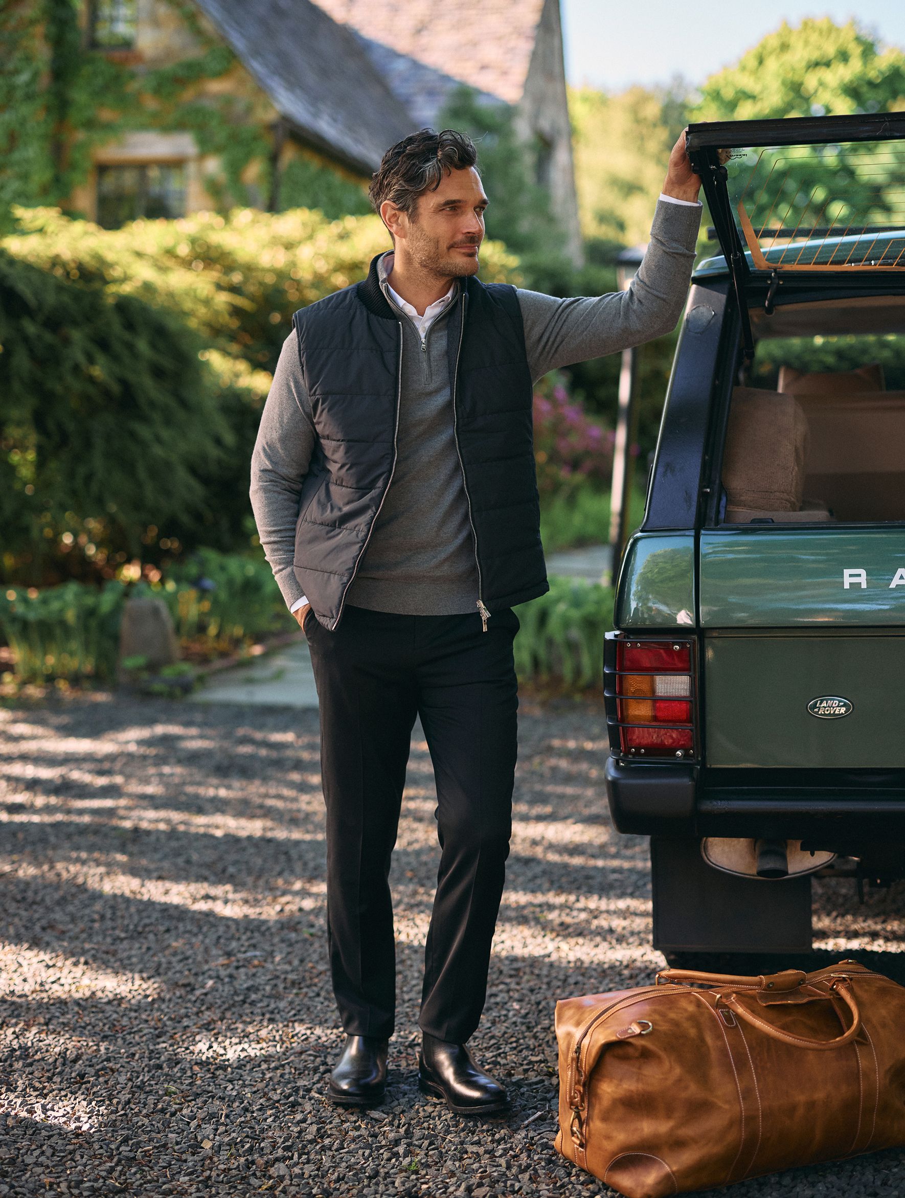 A man wearing a black puffy vest over a grey quarter-zip sweater, white collared shirt, and black trousers resting his arm on the open trunk of a green SUV next to a leather duffel bag.