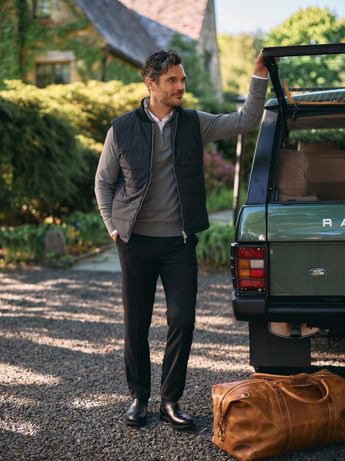 A man wearing a black puffy vest over a grey quarter-zip sweater, white collared shirt, and black trousers resting his arm on the open trunk of a green SUV next to a leather duffel bag.