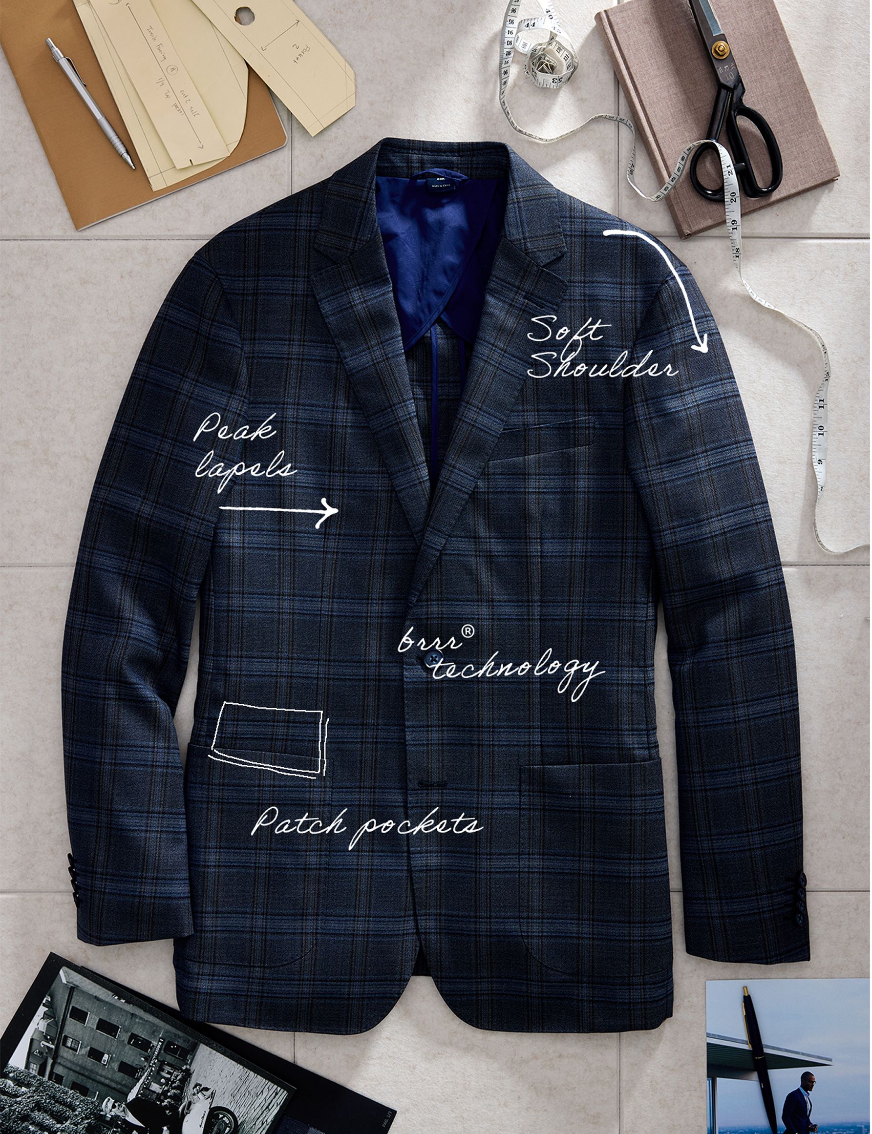 Product flat-lay of a dark blue plaid suit jacket with handwritten annotations identifying features like "Peak lapels", "Soft Shoulder", "brrr° technology", and "Patch pockets".