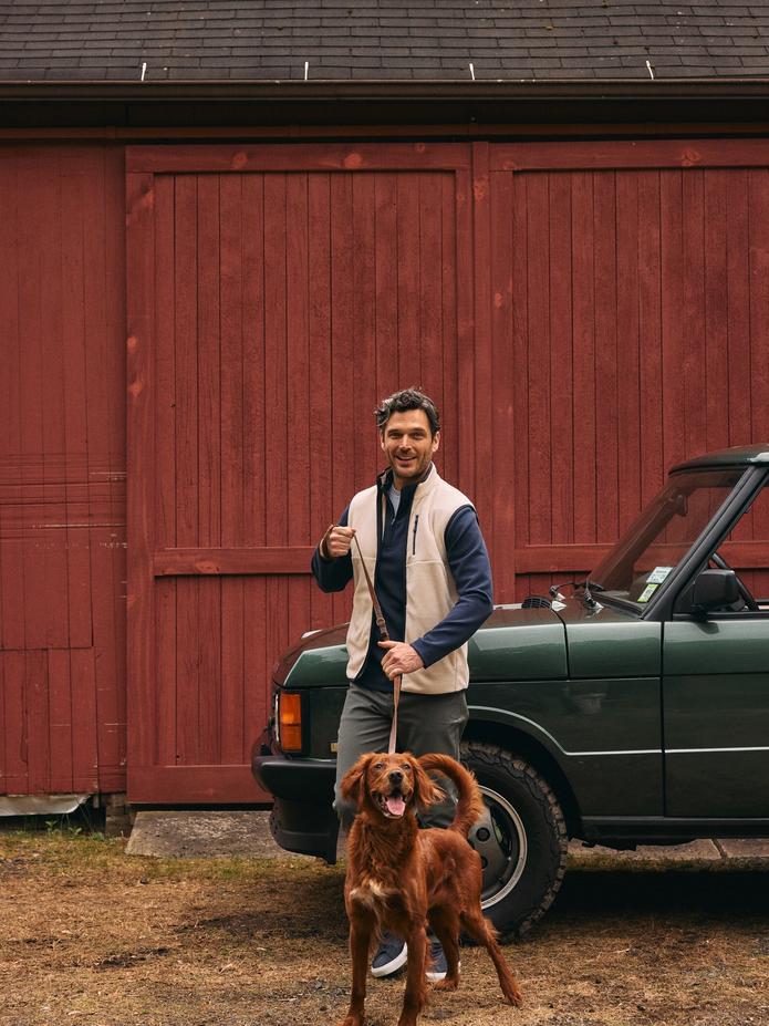 Man in a cream vest over a navy shirt walking an Irish setter in front of a red barn and a green Range Rover