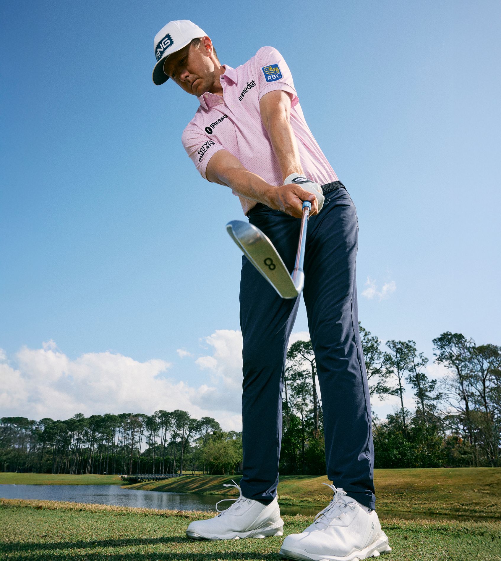 A golfer in a pink polo and navy performance pants follows through on an iron shot beside a fairway lake.