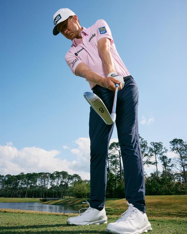 A golfer in a pink polo and navy performance pants follows through on an iron shot beside a fairway lake.