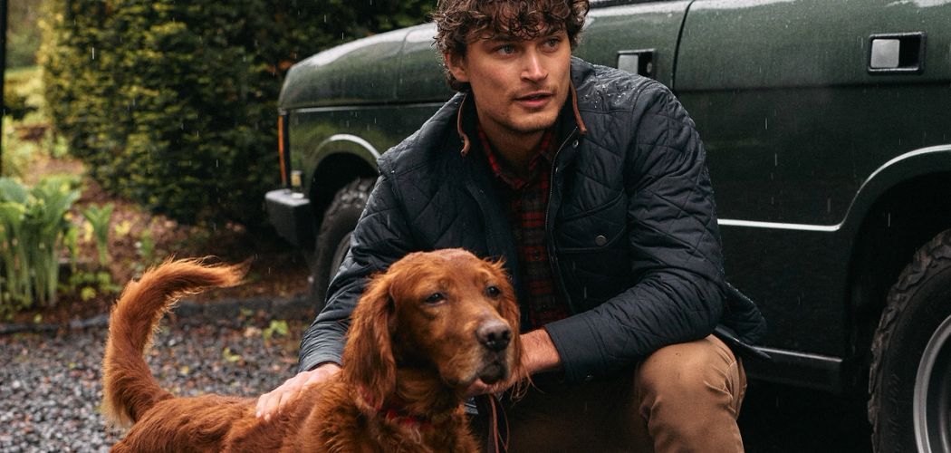 Man in a navy quilted jacket crouching beside a red setter in front of a dark green vintage SUV