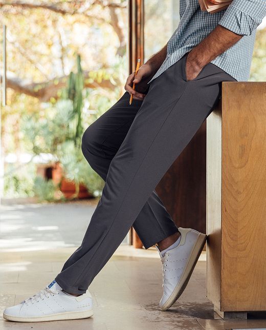 A man leans against a wooden counter in gray performance pants and white sneakers.