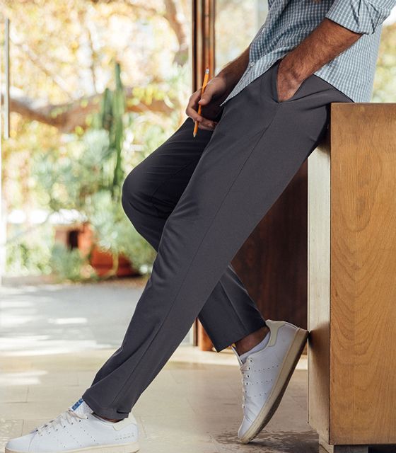 A man leans against a wooden counter in gray performance pants and white sneakers.