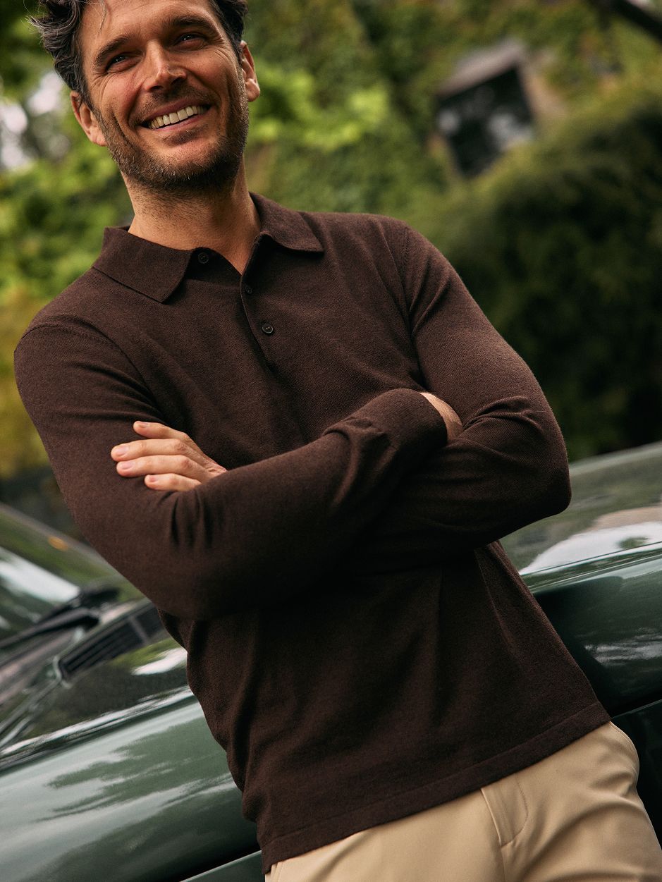 A man with dark wavy hair smiling with arms crossed while leaning on a green vehicle, wearing a dark brown long-sleeve knit polo shirt and tan pants.