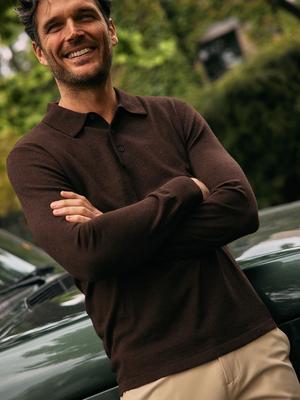 A man with dark wavy hair smiling with arms crossed while leaning on a green vehicle, wearing a dark brown long-sleeve knit polo shirt and tan pants.