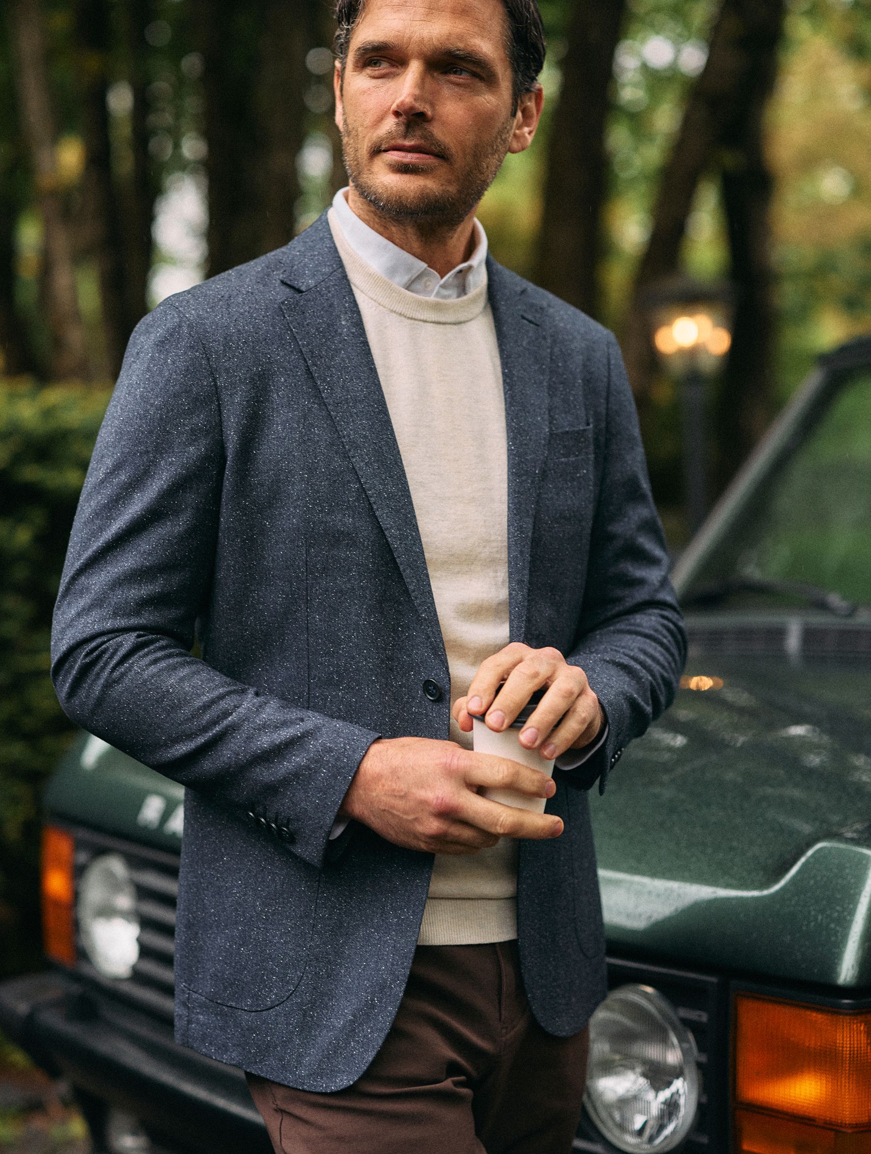 A man holding a coffee cup while standing in front of a green SUV, wearing a grey textured blazer, off-white sweater, collared shirt, and brown pants.
