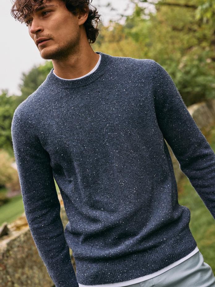 A man wearing a blue speckled crewneck sweater over a white t-shirt standing outdoors.