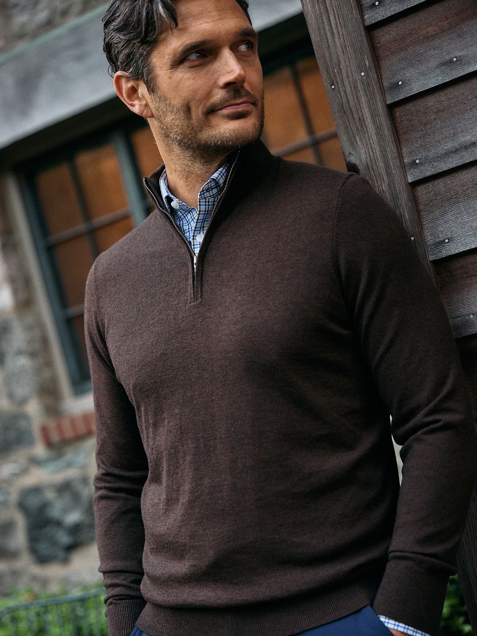 A man wearing a dark brown quarter-zip sweater over a blue plaid collared shirt, leaning against a wooden doorframe.
