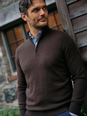 A man wearing a dark brown quarter-zip sweater over a blue plaid collared shirt, leaning against a wooden doorframe.